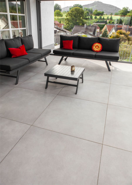 Eine Terrasse mit dunkelgrauen Polstermöbeln mit roten Kissen und großformatigen, hellgrauen Bodenfliesen. Im Hintergrund ist eine grüne Aussicht in die Natur und Berge