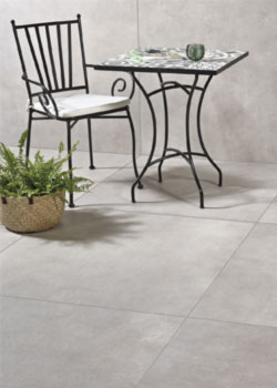 Graue Terrassenplatten im Material "Berlin Grey" mit einem schwarzen Tisch und Stuhl. daneben steht ein Farn im Bastkorb