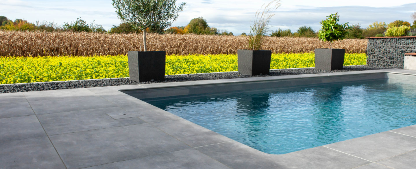 Blick auf einen Pool mit grauen Poolrandplatten vor einem gelben Rapsfeld