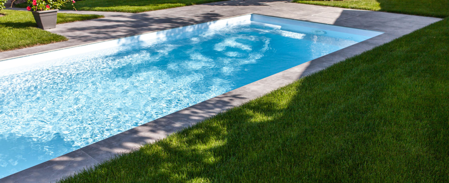 Pool mit hellblauem Wasser, Poolrandplatten in grauer Betonoptik und ein saftig grüner Rasen