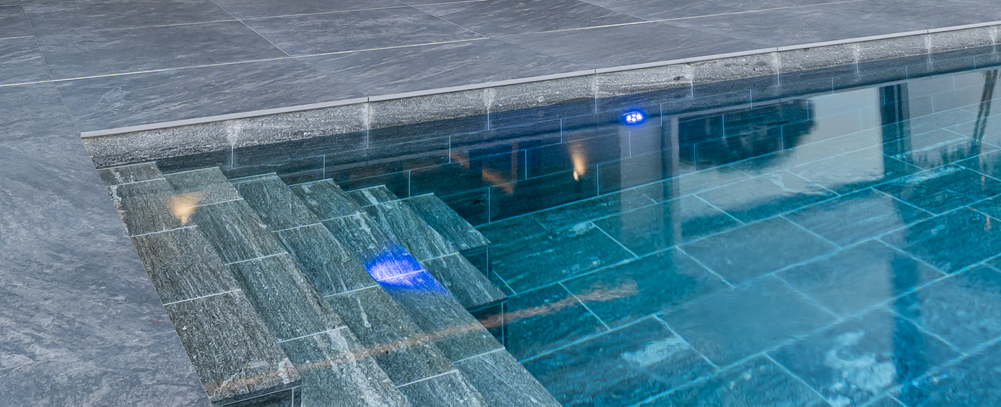 Belagstreppe die als Einstieg in einen grau gefliesten Pool führt aus Naturstein Biasca Gneis