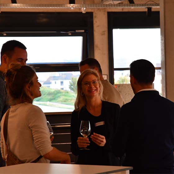 linda-wein-rasterbild Eine Gruppe von Menschen steht im Kreis vor einem Fenster und trinken Wein.