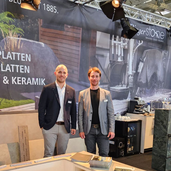 Simeon_Lucas_Messestand Zwei Personen lächeln in die Kamera und stehen auf einem Messestand.